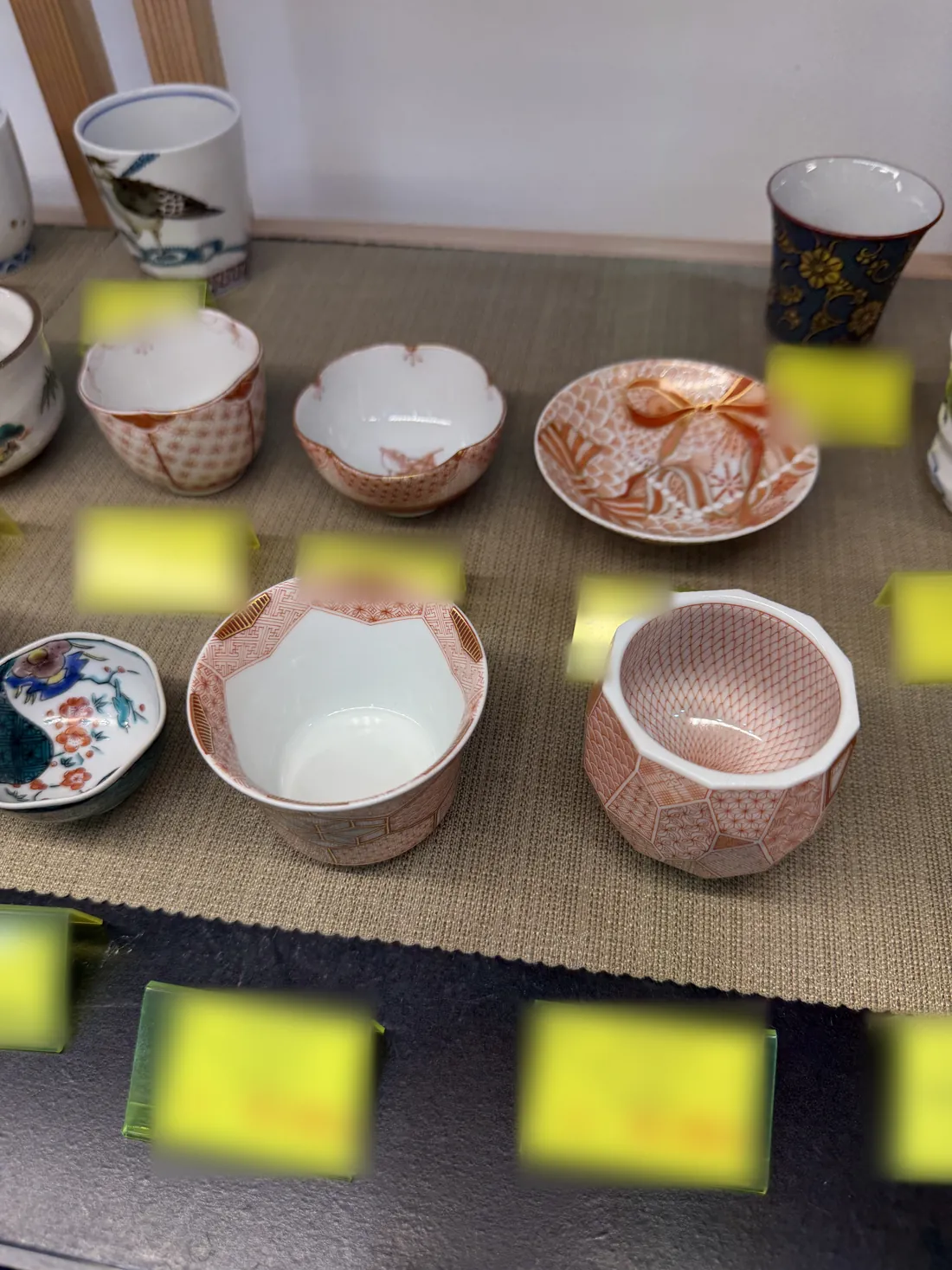 Red-painted Kutani ware sake cups and a small plate with fine red patterns on the shelf, with the price tags blurred