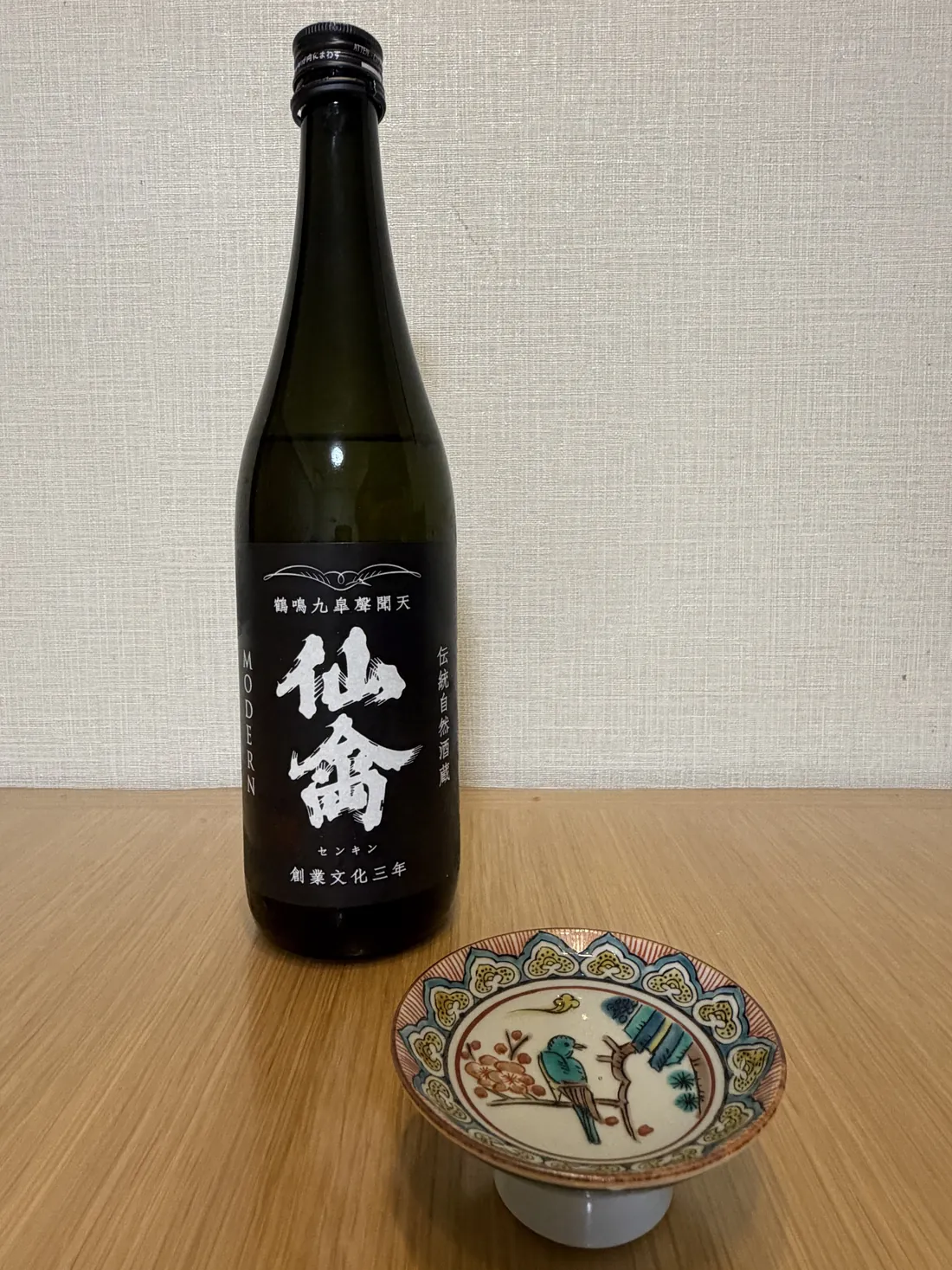 A black Senkin Modern sake bottle beside the Kutani sakazuki on a wooden table