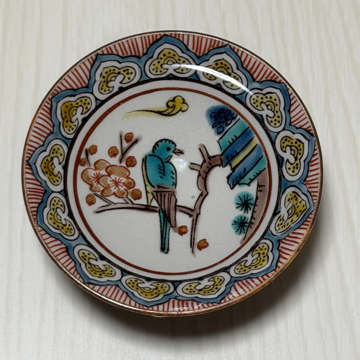 Kutani ware sakazuki seen directly from above, showing a turquoise bird on a flowering branch inside a border of red lines and yellow and blue-green shapes